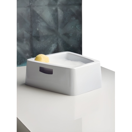 Fußbadewanne mit Schaum und Schwamm auf weißer Oberfläche vor grauer Wand, ideal für entspannende Fußpflege.